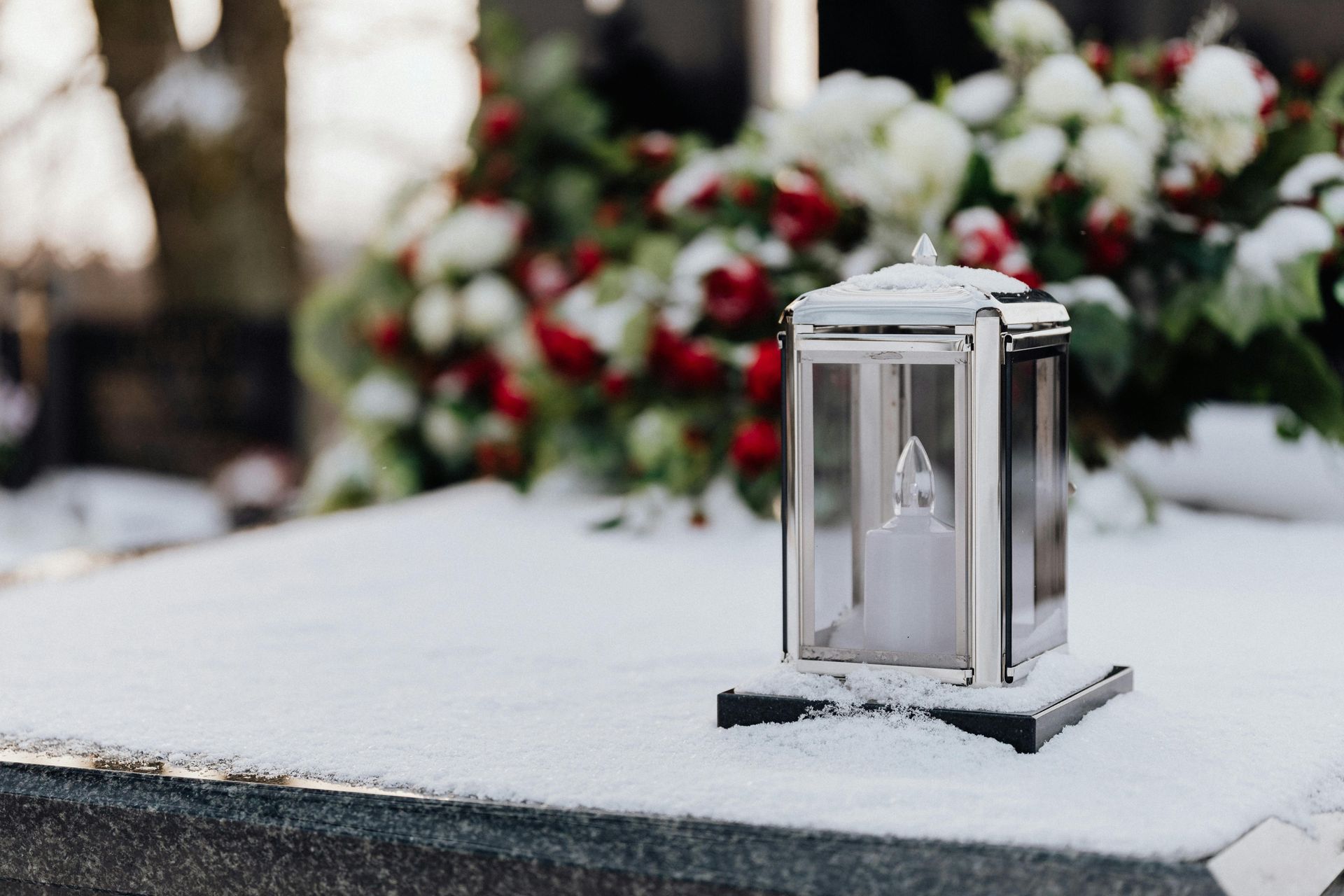 A lit candle lantern sits on a snow-covered tombstone, with red and white flowers in the background.