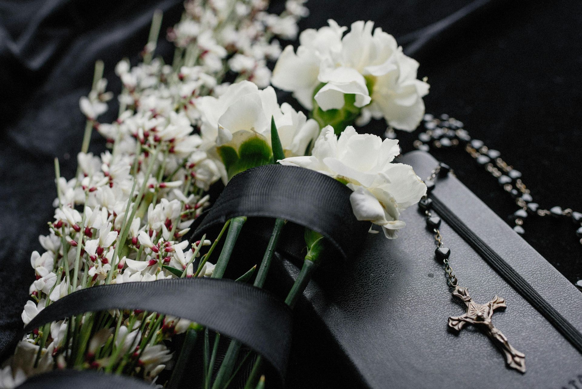 White flowers, black ribbon, rosary, and a crucifix on a dark surface, likely a memorial or mourning scene.