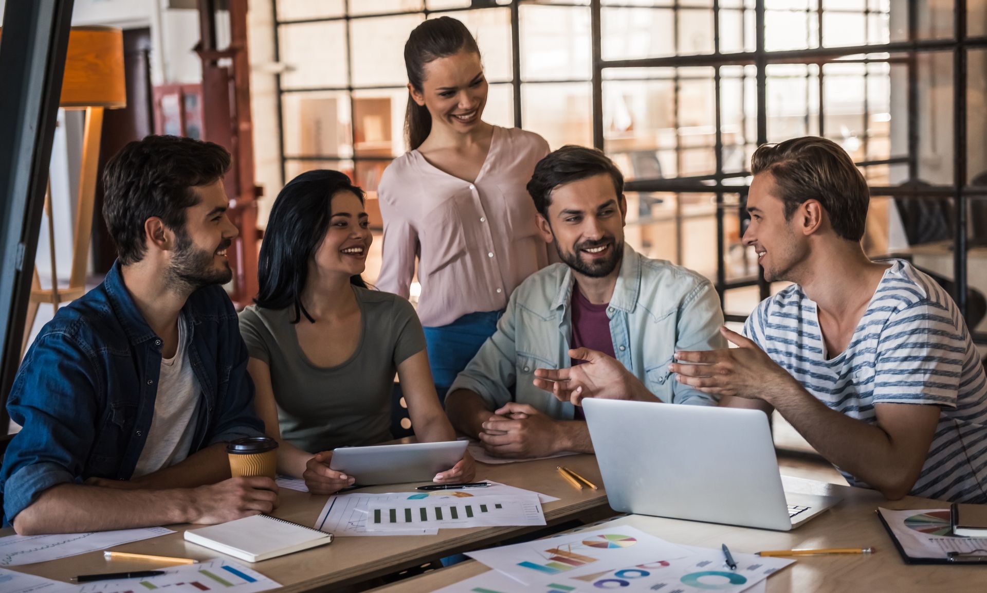 Group of people collaborating around a table with a laptop, documents, and coffee, in a modern office space.