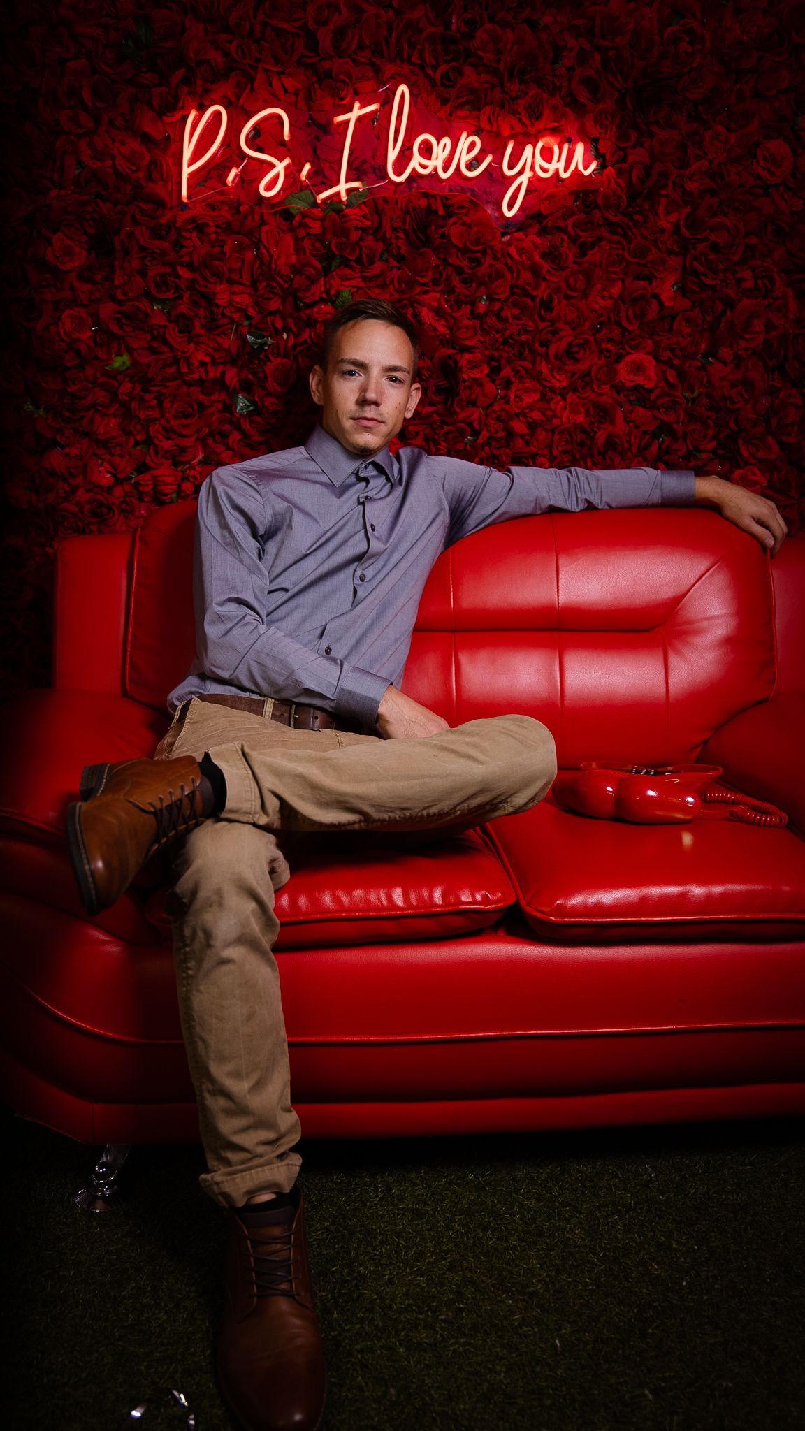 A man is sitting on a red couch in front of a wall of red roses.