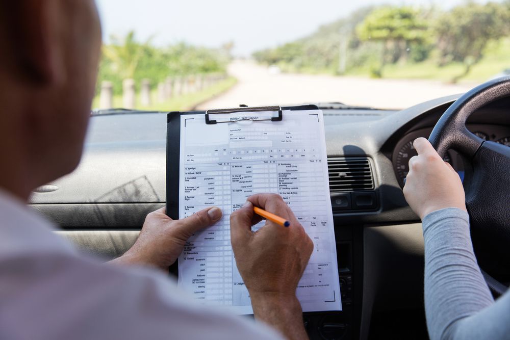 Driving instructor in car writing on a clipboard, pointing to the steering wheel, road in the distance. — Des’s Driving School in Callala Bay, NSW