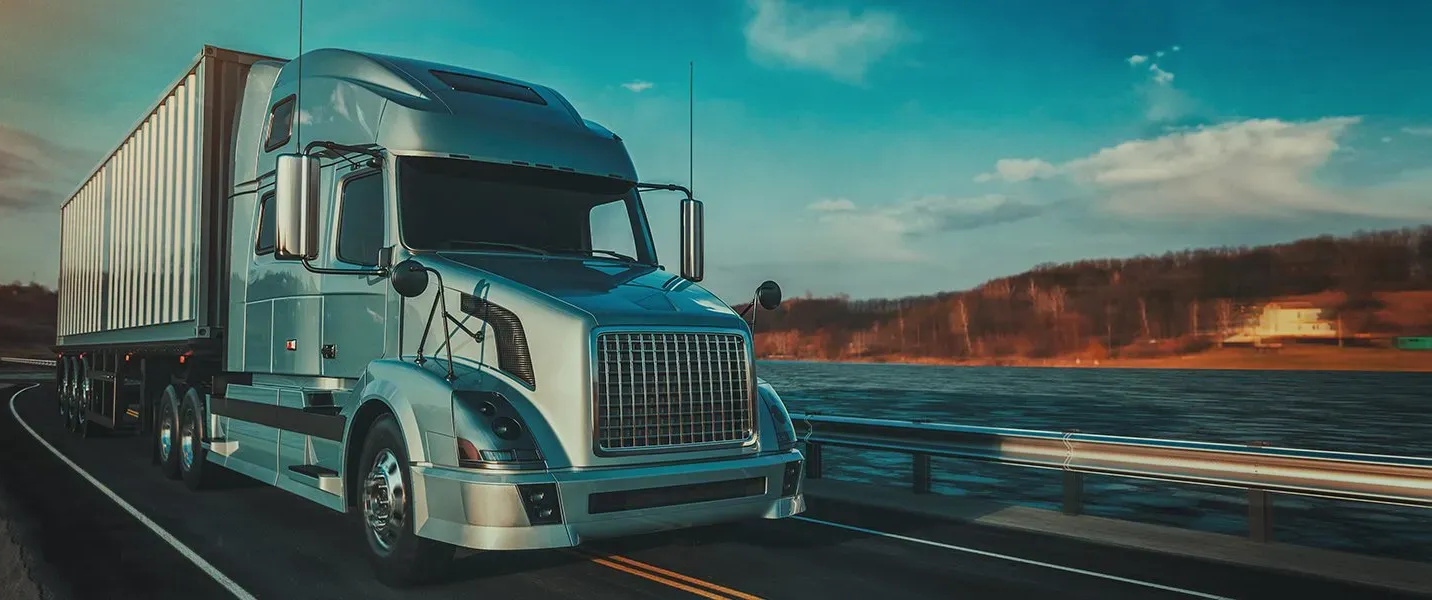 A silver semi-truck drives on a road by a lake. The sky is blue and the landscape is visible. — Des’s Driving School in Callala Bay, NSW