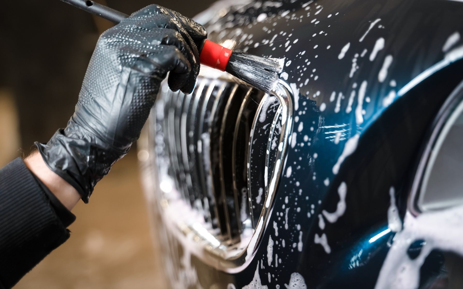 Gloved hand using a brush to clean a car grill with soapy water. Black car.