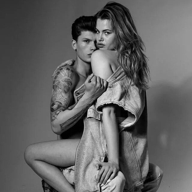 Man with arm tattoos embraces woman; both wear denim, black and white.