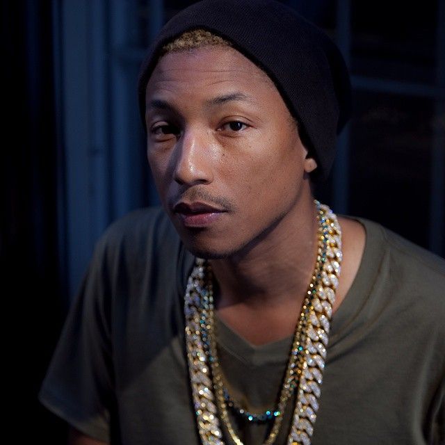 Pharrell Williams in a black beanie and multiple gold chains, looking at the camera.