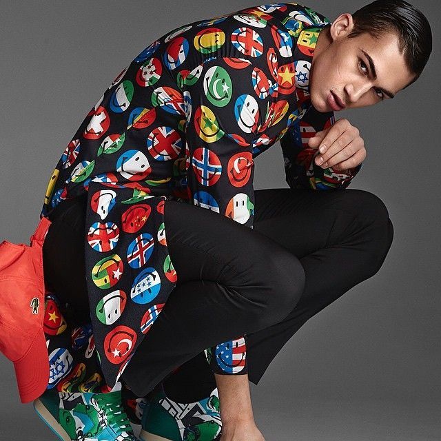 Man squatting, wearing a black shirt with flag smiley faces, black pants, and colorful sneakers.