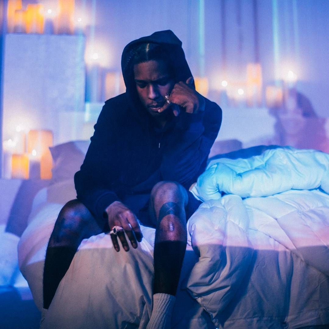 A$AP Rocky sitting on a bed, wearing a hoodie and shorts, looking down with a pensive expression.