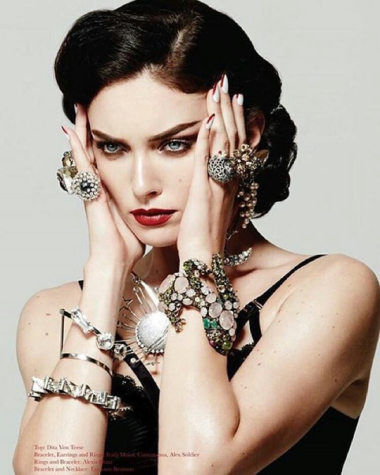 Woman with dark hair, wearing many rings and bracelets, touches her face. She has red lipstick.