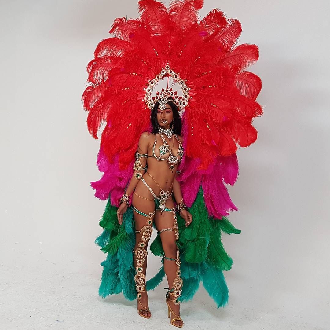 Woman in vibrant carnival costume, large red feather headdress, jewel-covered body suit, white background.
