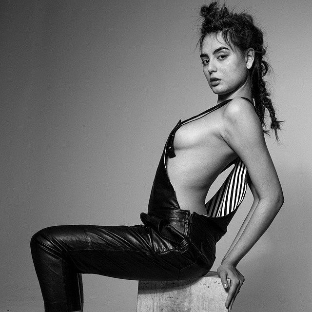 Woman in black leather overalls, posing dramatically, seated on a block.