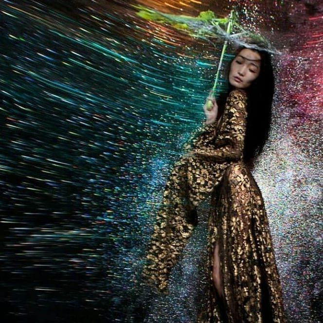 Woman in gold gown holding umbrella in colorful, swirling water.