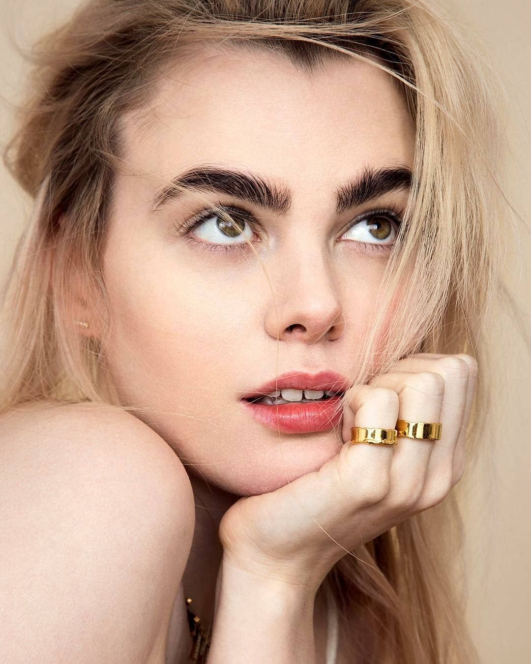 Blonde woman with thick eyebrows gazes upward, resting chin on gold rings, pink lips, beige background.