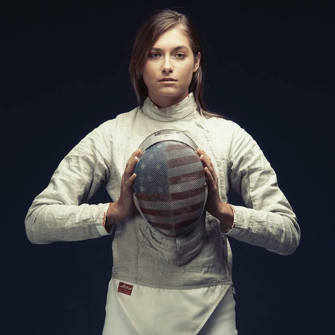 Female fencer in uniform holding her mask, arms outstretched, against a dark background.