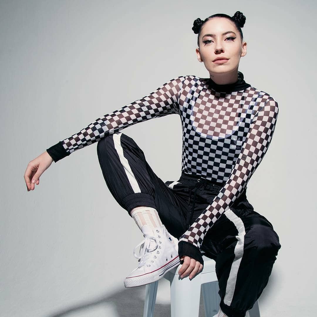 Woman in checkered top, seated on a stool, with space buns, black pants, and white sneakers.