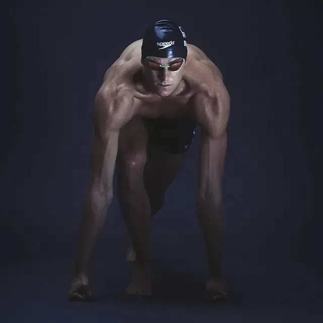 Swimmer in starting position, wearing cap and goggles, dark background.