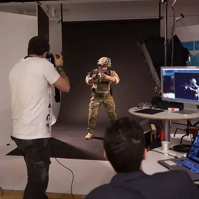Photographer taking a picture of soldier in camouflage with a gun against a black backdrop in a studio.