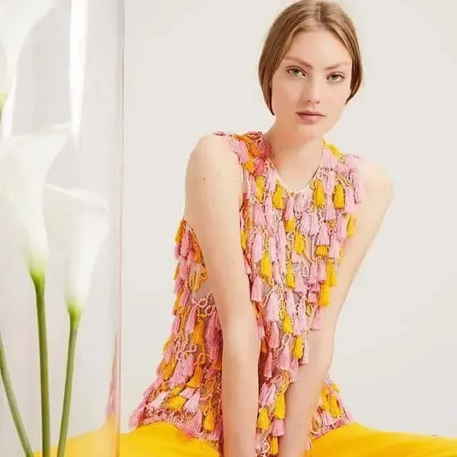 Woman wearing a pink and yellow tassel top, posing indoors with calla lilies.