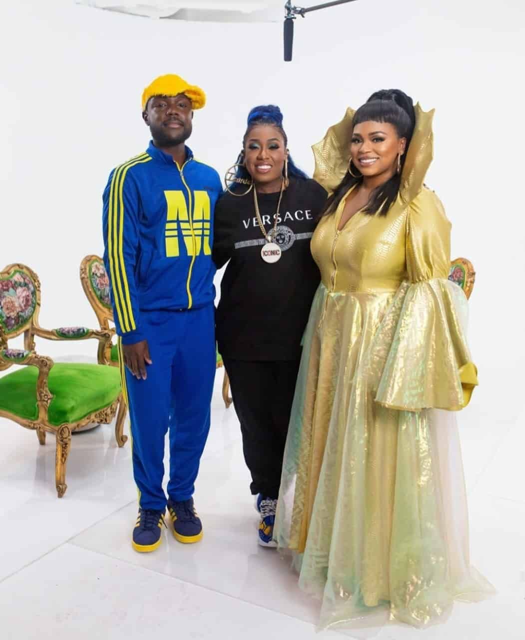 Three people pose against a white backdrop. A man in blue sweatsuit, a woman in black, and a woman in a gold gown.