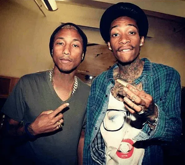 Pharrell Williams and Wiz Khalifa posing, Pharrell pointing, Wiz smoking, indoors.
