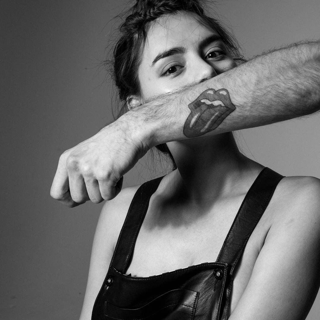 Woman's face partially covered by arm with Rolling Stones tattoo, wearing black overalls.