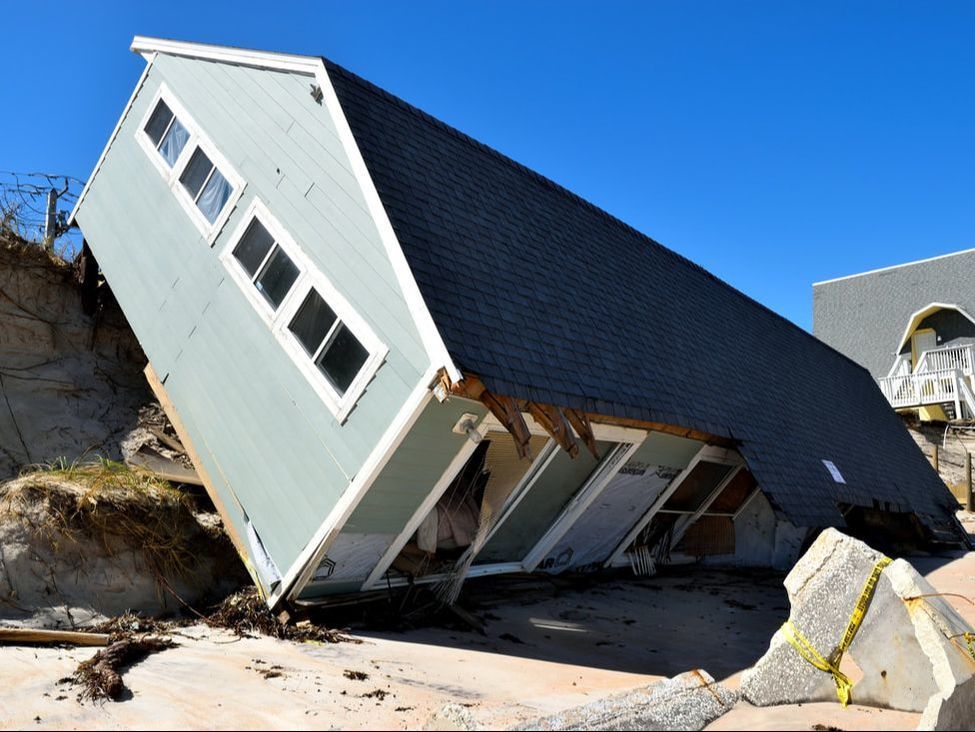 House damaged and tilted on a sandy beach after a storm, with a blue sky background.