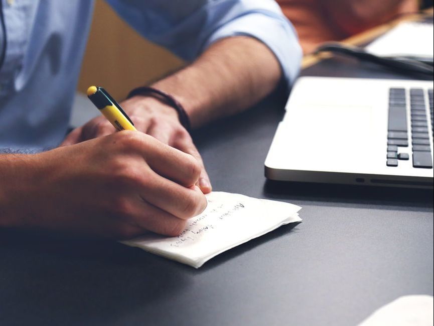 Person writes on a small white notepad with a yellow and black pen near a laptop on a dark desk.