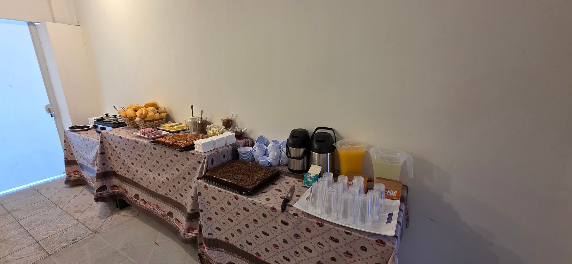 Mesa de buffet posta com comidas e bebidas, em ambiente interno. Toalha de mesa marrom com bolinhas. Inclui café, suco e doces.