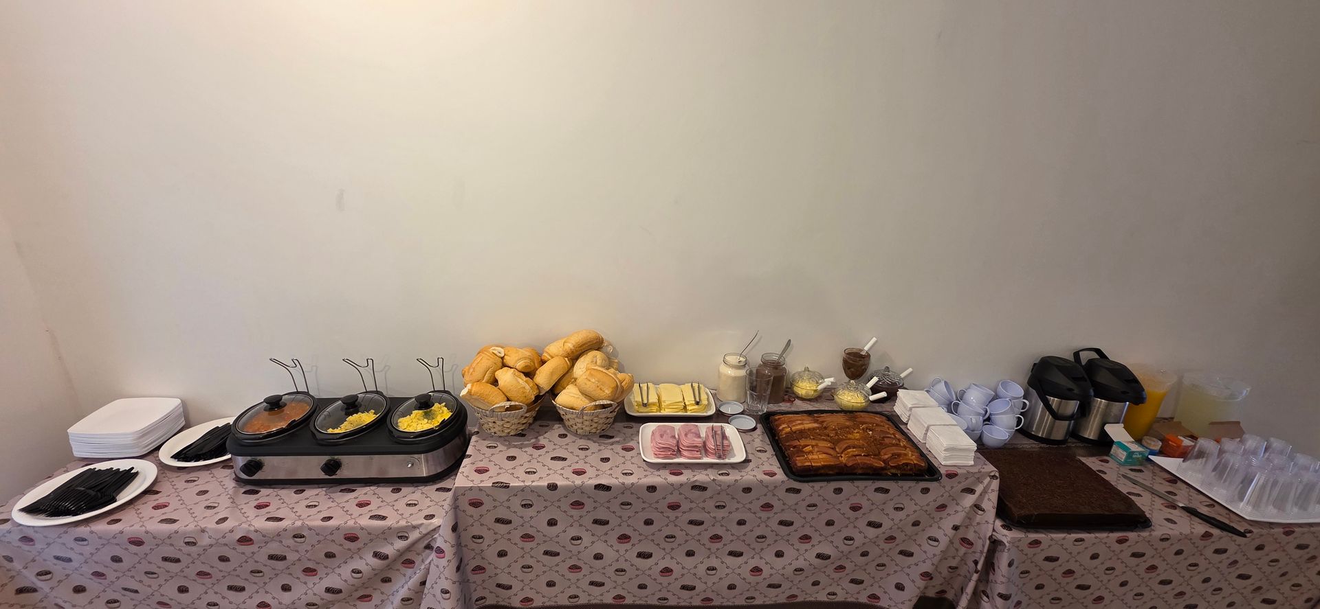 Buffet de café da manhã com pão, doces, presunto e chapa elétrica em uma mesa com toalha de mesa estampada.