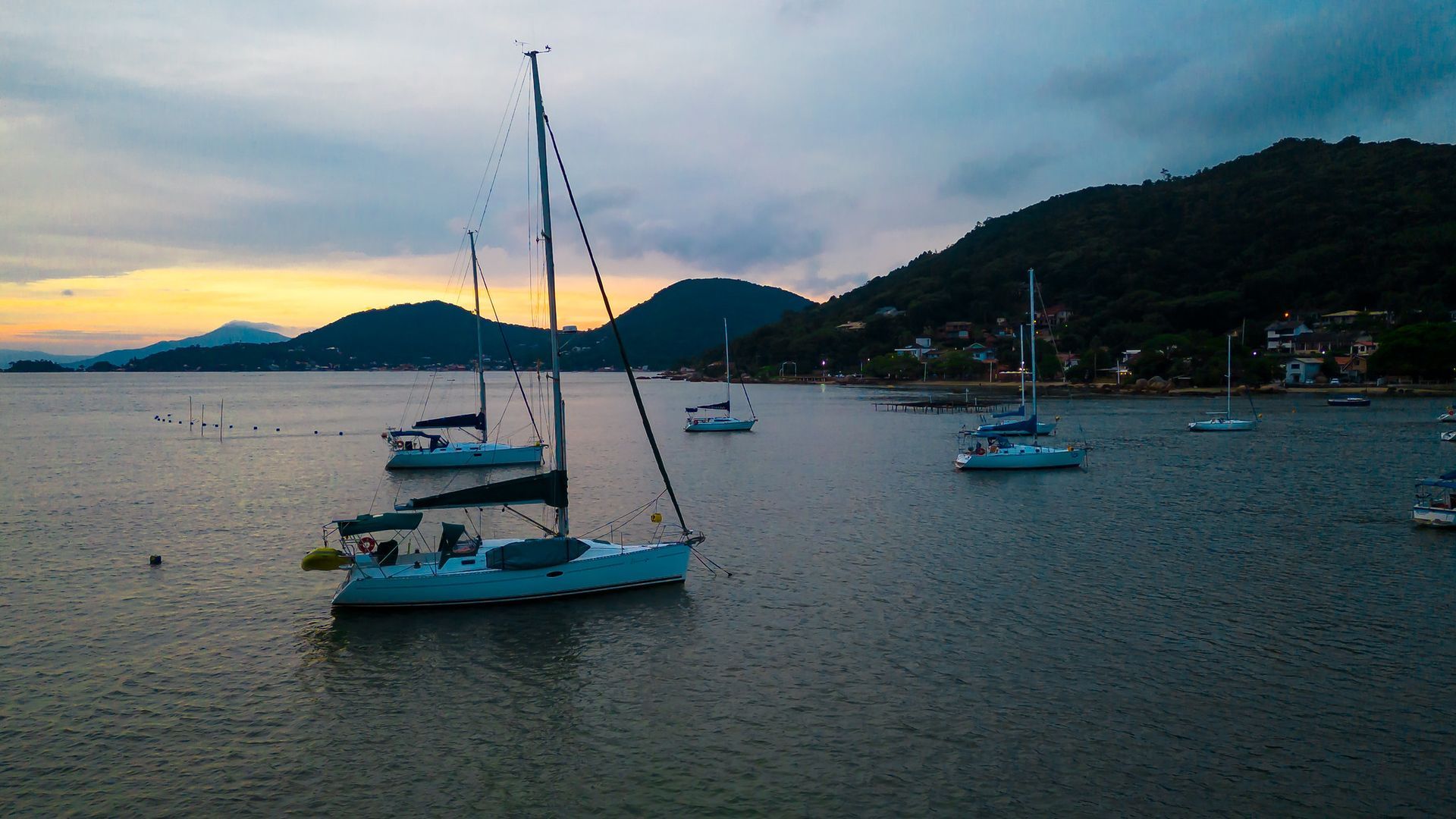 Veleiros em águas calmas ao entardecer, com um cenário montanhoso e céu nublado.