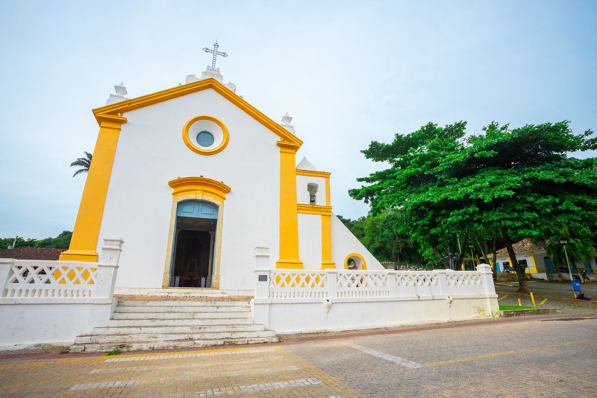 Igreja branca com detalhes em amarelo, degraus e uma grande árvore verde ao lado.