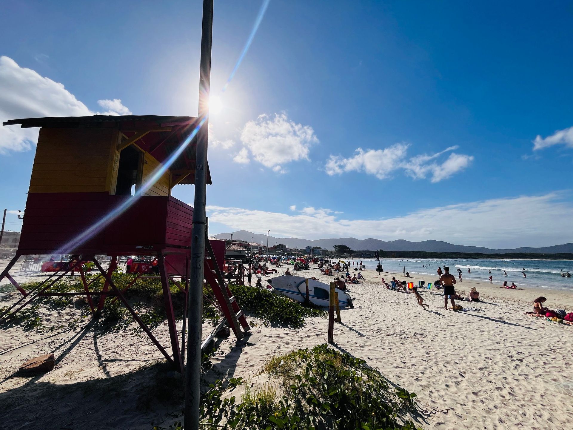 Cena de praia com posto de salva-vidas, céu ensolarado e pessoas relaxando na areia.
