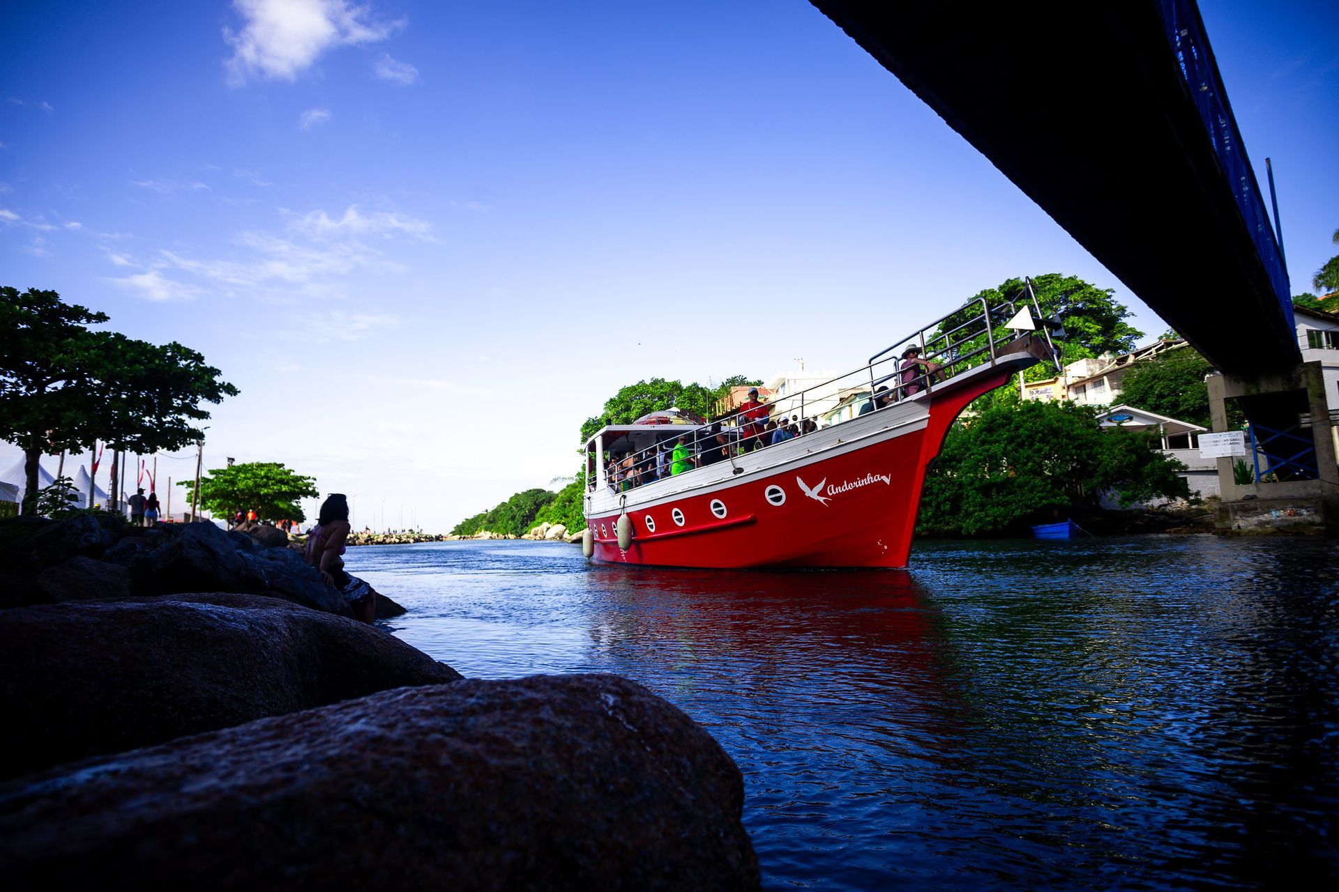 Barco vermelho navegando na água, perto de uma costa rochosa e sob uma ponte, em um dia ensolarado.