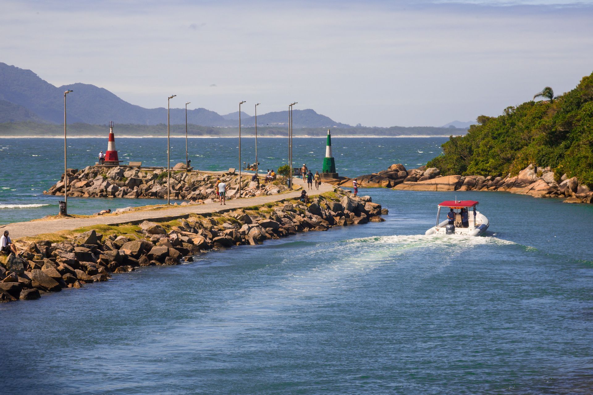 Barco navegando por um canal entre quebra-mares rochosos com faróis vermelhos e verdes sob um céu azul, com montanhas ao longe.