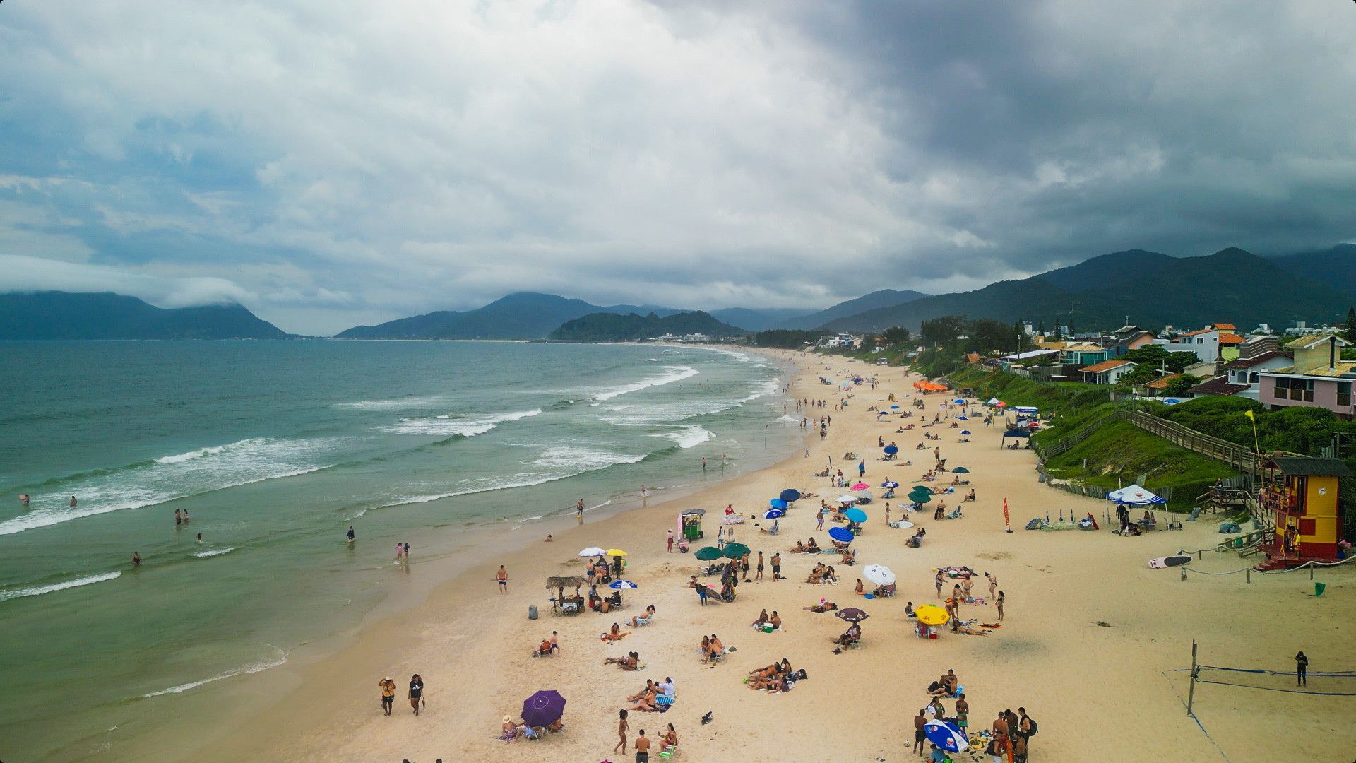 Cena de praia com pessoas, ondas do oceano e céu nublado.