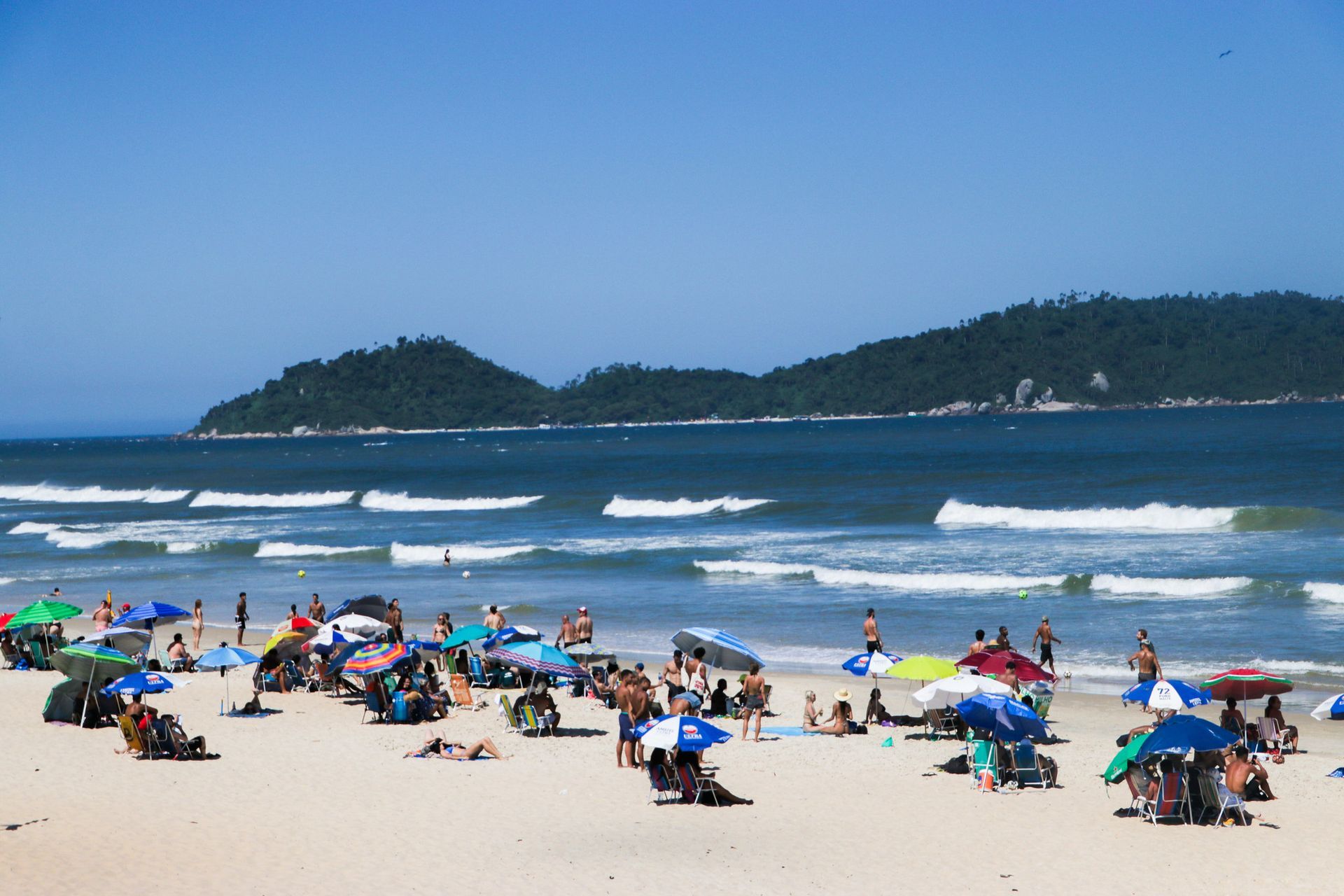 Cena de praia com pessoas sob guarda-sóis, ondas do oceano e colinas em um dia ensolarado.