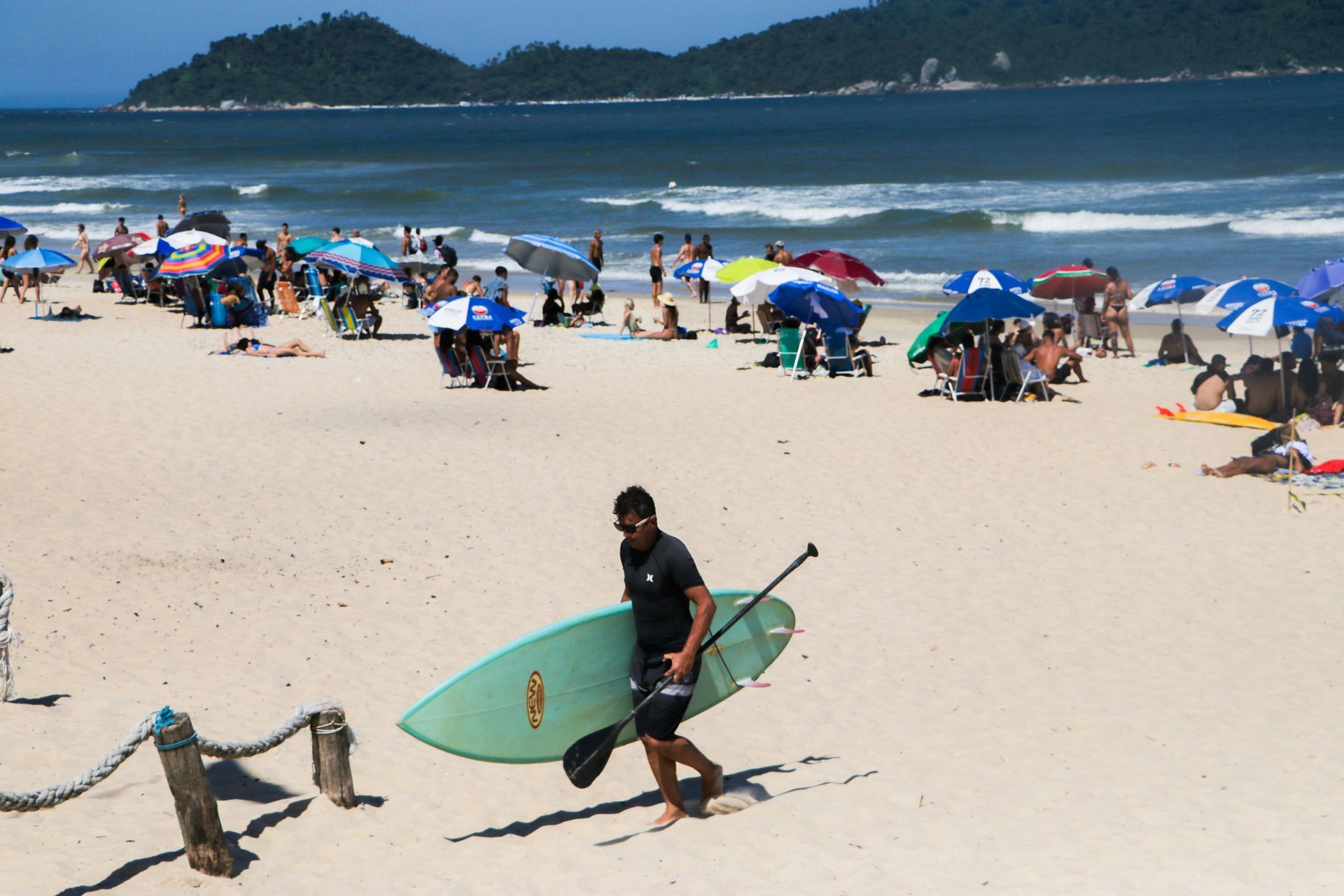 Homem carregando uma prancha de surfe azul-clara em uma praia lotada com guarda-sóis e o oceano ao fundo.