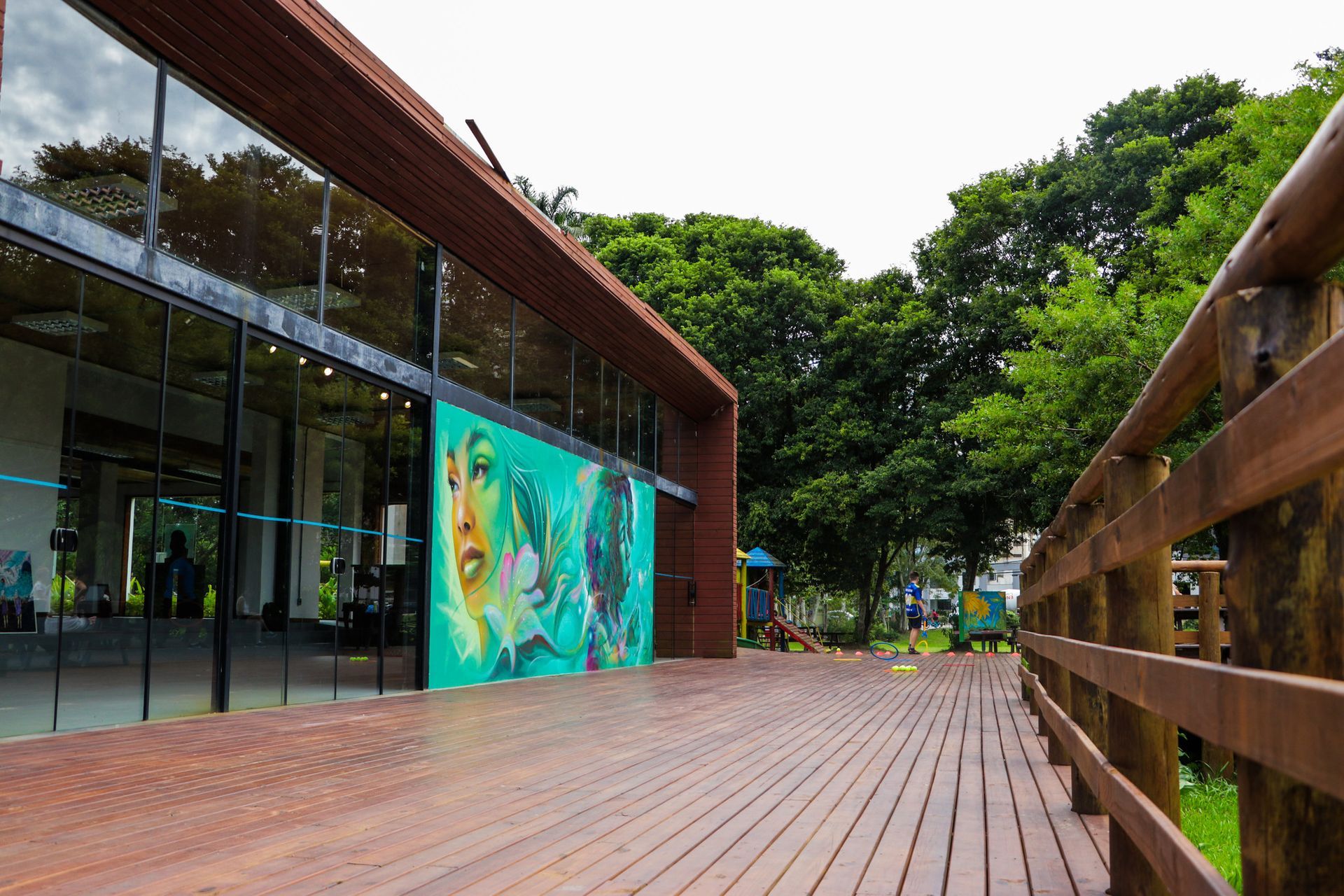 Passarela de madeira ao lado de um prédio moderno com grandes janelas e um mural turquesa. Árvores verdejantes ao fundo.