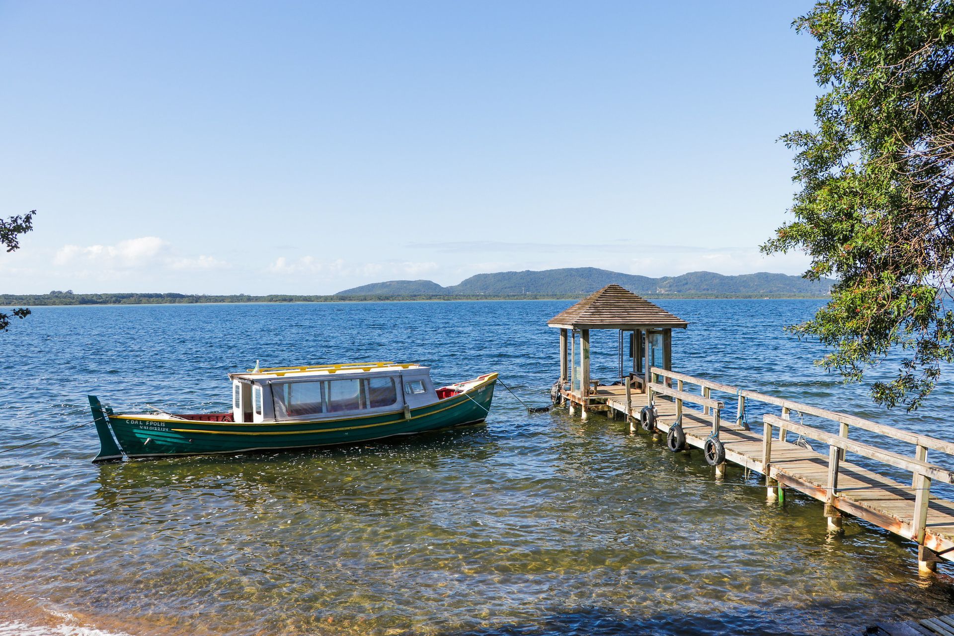 Barco verde próximo a um píer de madeira em um lago sob um céu azul, com montanhas ao longe.