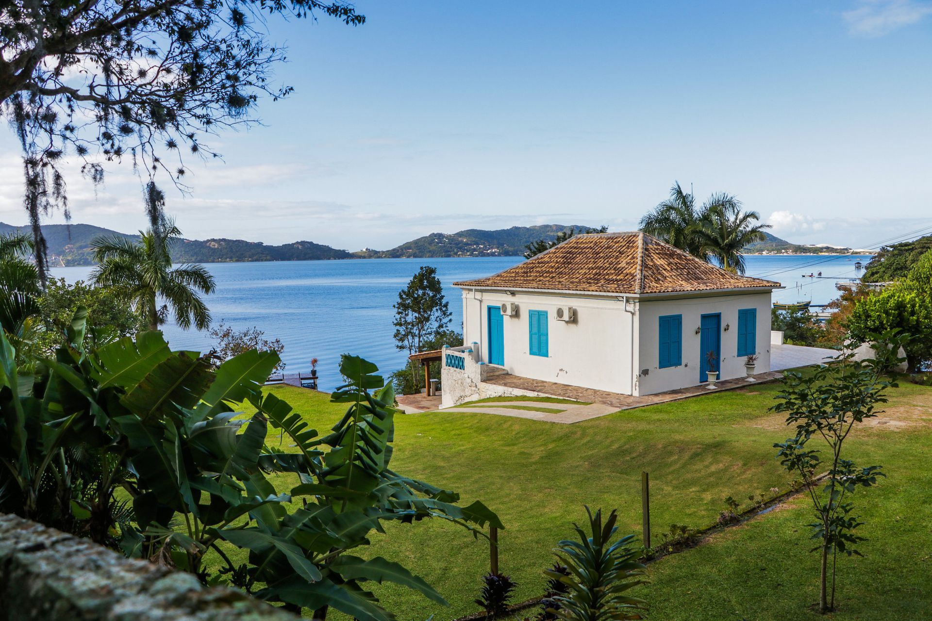 Casa branca com venezianas azuis com vista para um oceano azul. Grama verde e árvores em primeiro plano.