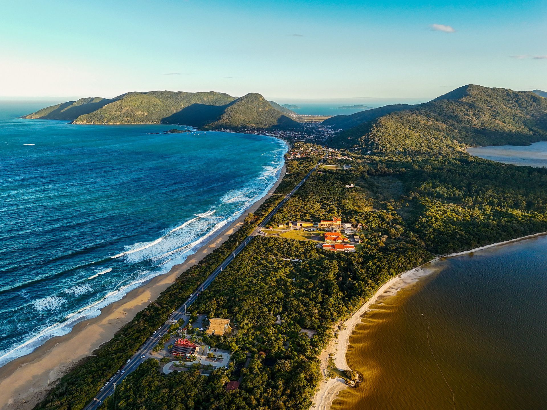 Vista aérea de uma paisagem costeira com praia, oceano, lago, árvores verdejantes e colinas distantes sob um céu azul.