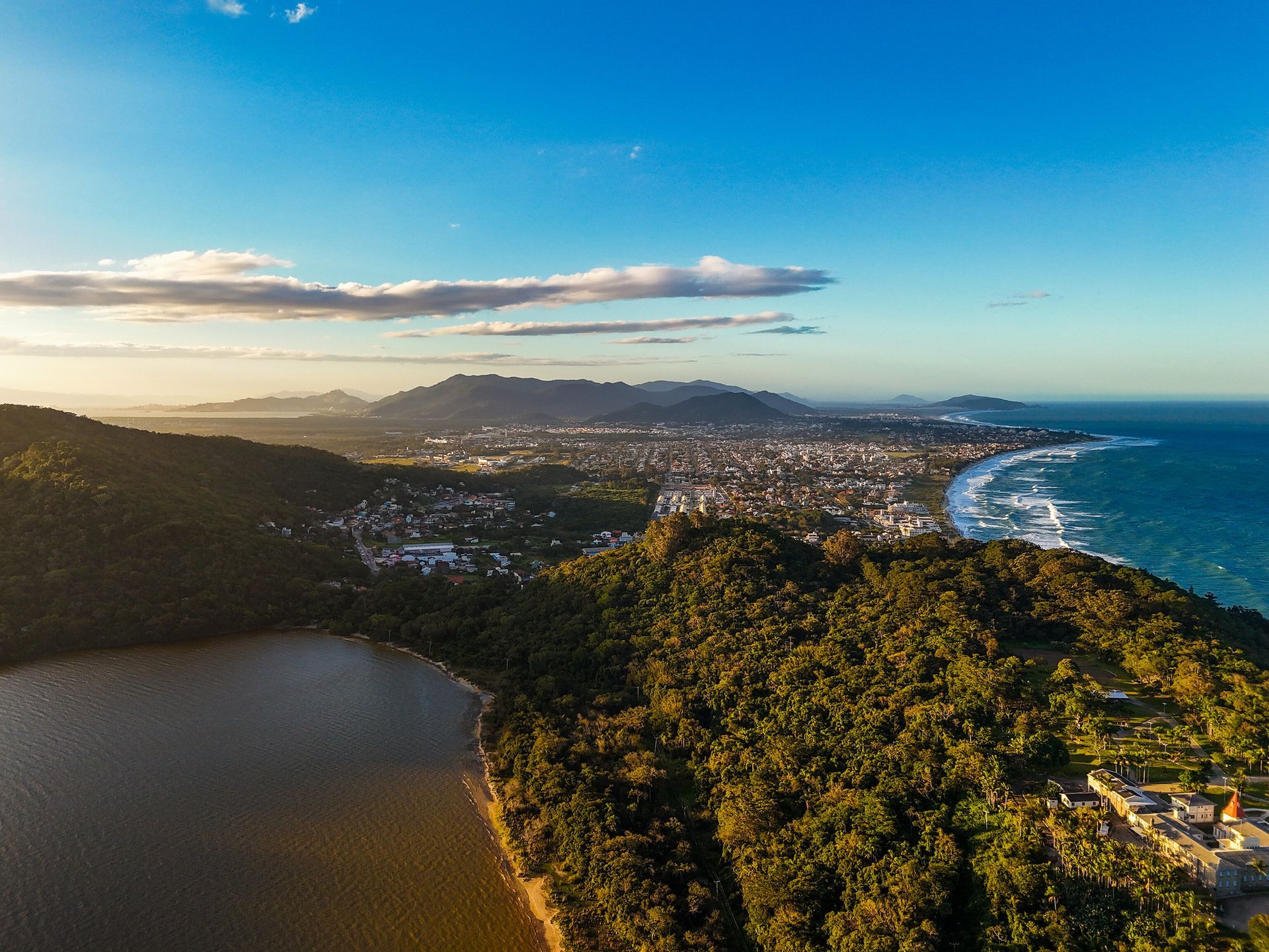 Vista aérea de uma cidade costeira com uma floresta e um lago em primeiro plano e o oceano à direita, sob um céu azul.