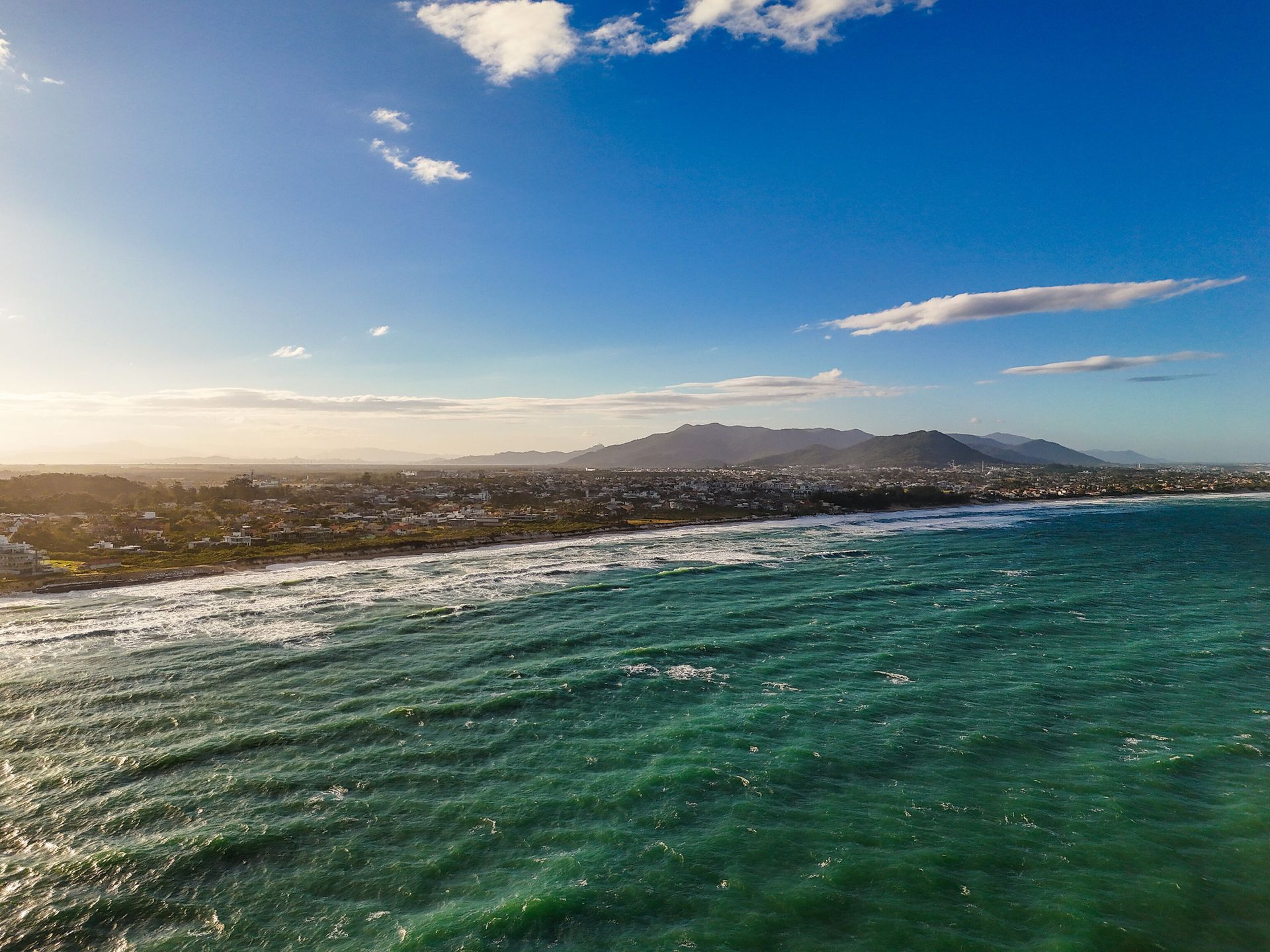 Ondas do oceano quebrando na praia, prédios da cidade e montanhas ao longe sob um céu azul.