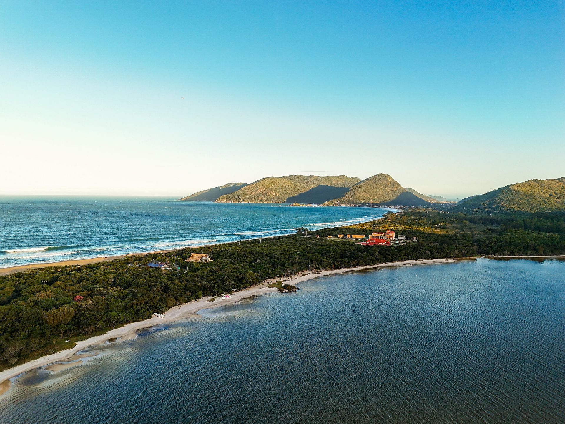 Vista aérea de uma restinga que se projeta para o mar, com uma praia de um lado e terreno coberto de vegetação do outro.