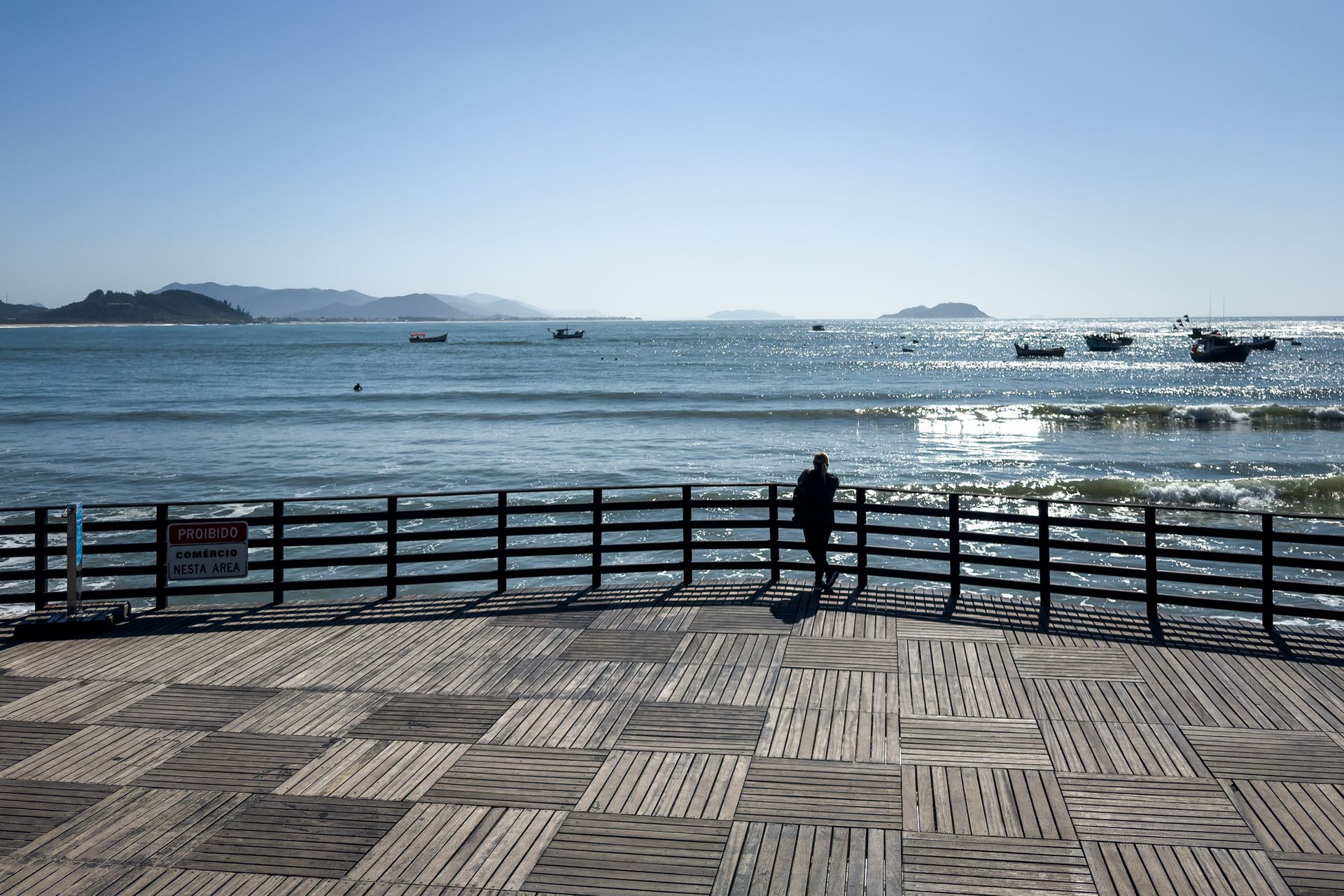 Pessoa em um deck de madeira olhando para o oceano com barcos, céu azul.
