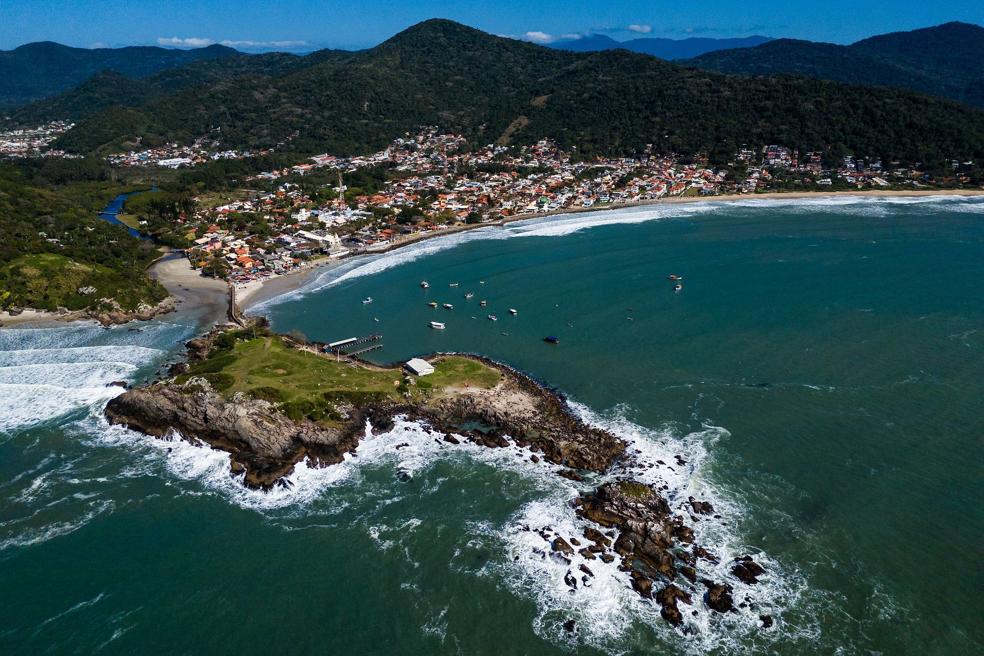 Cidade costeira com vista para o oceano, com uma ilha rochosa em primeiro plano. Montanhas e edifícios visíveis.