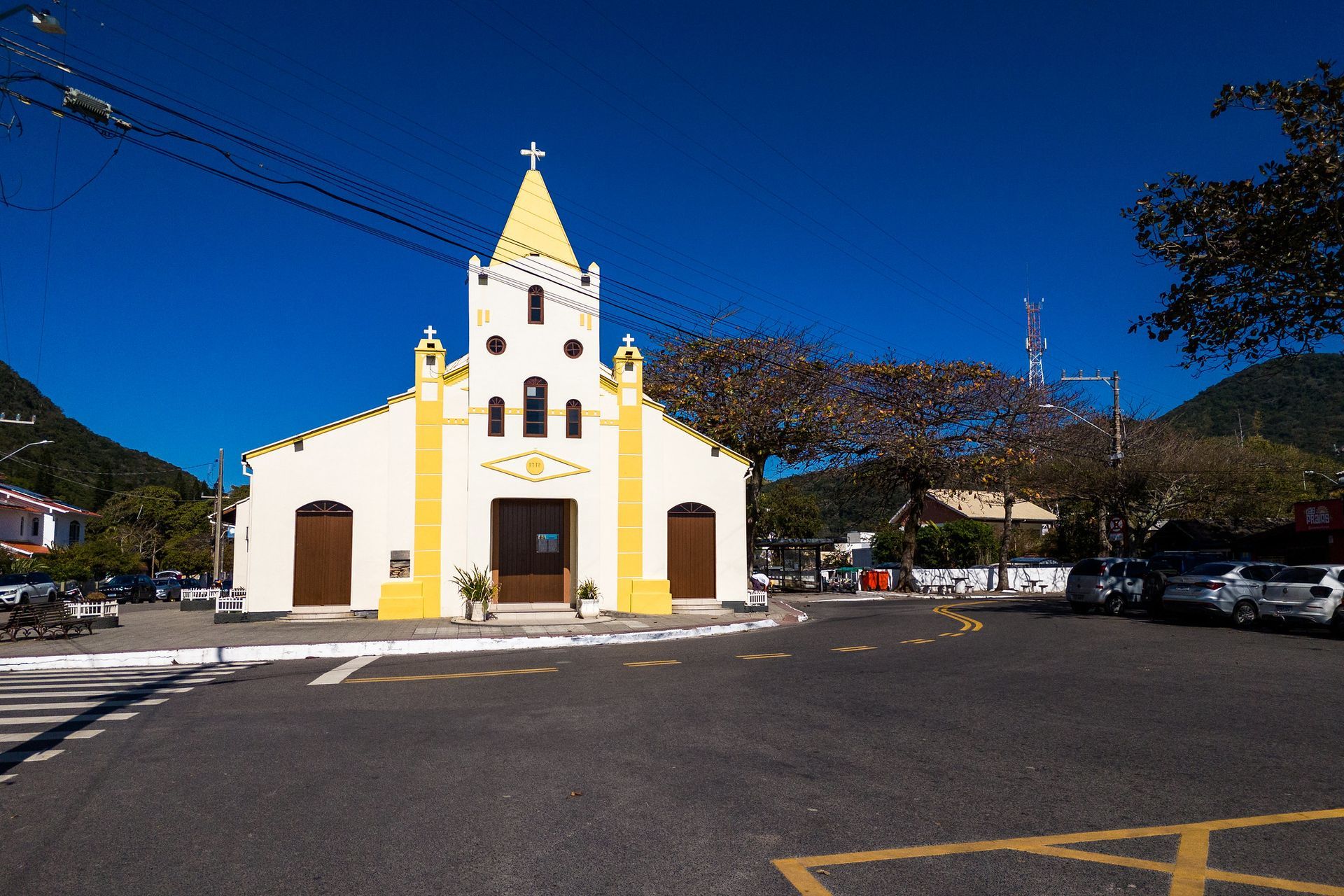 Igreja amarela com telhado pontiagudo e cruz, contra um céu azul claro. Rua em primeiro plano.