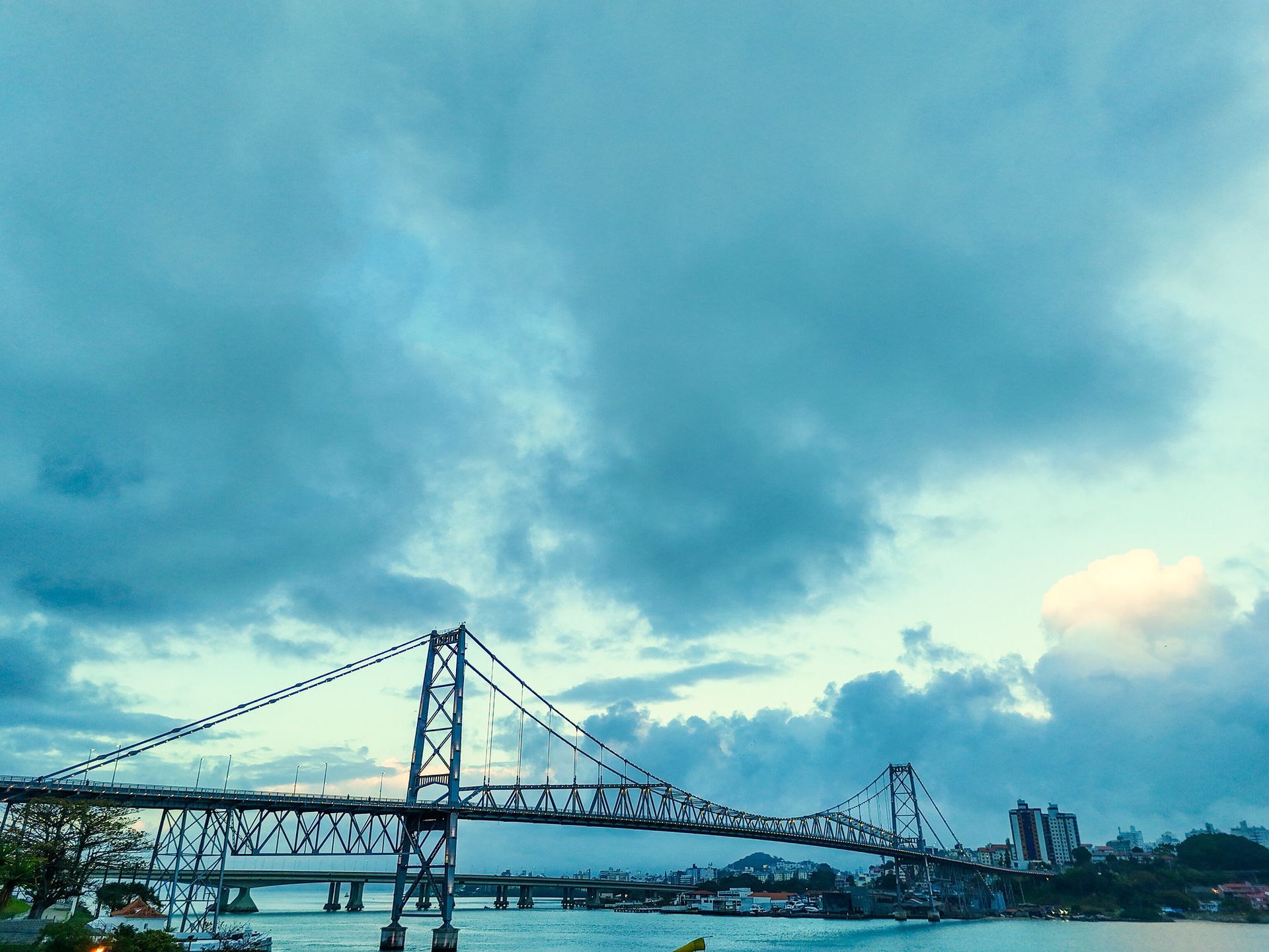 Ponte suspensa sobre a água sob um céu nublado de tons azulados. Prédios visíveis à direita.