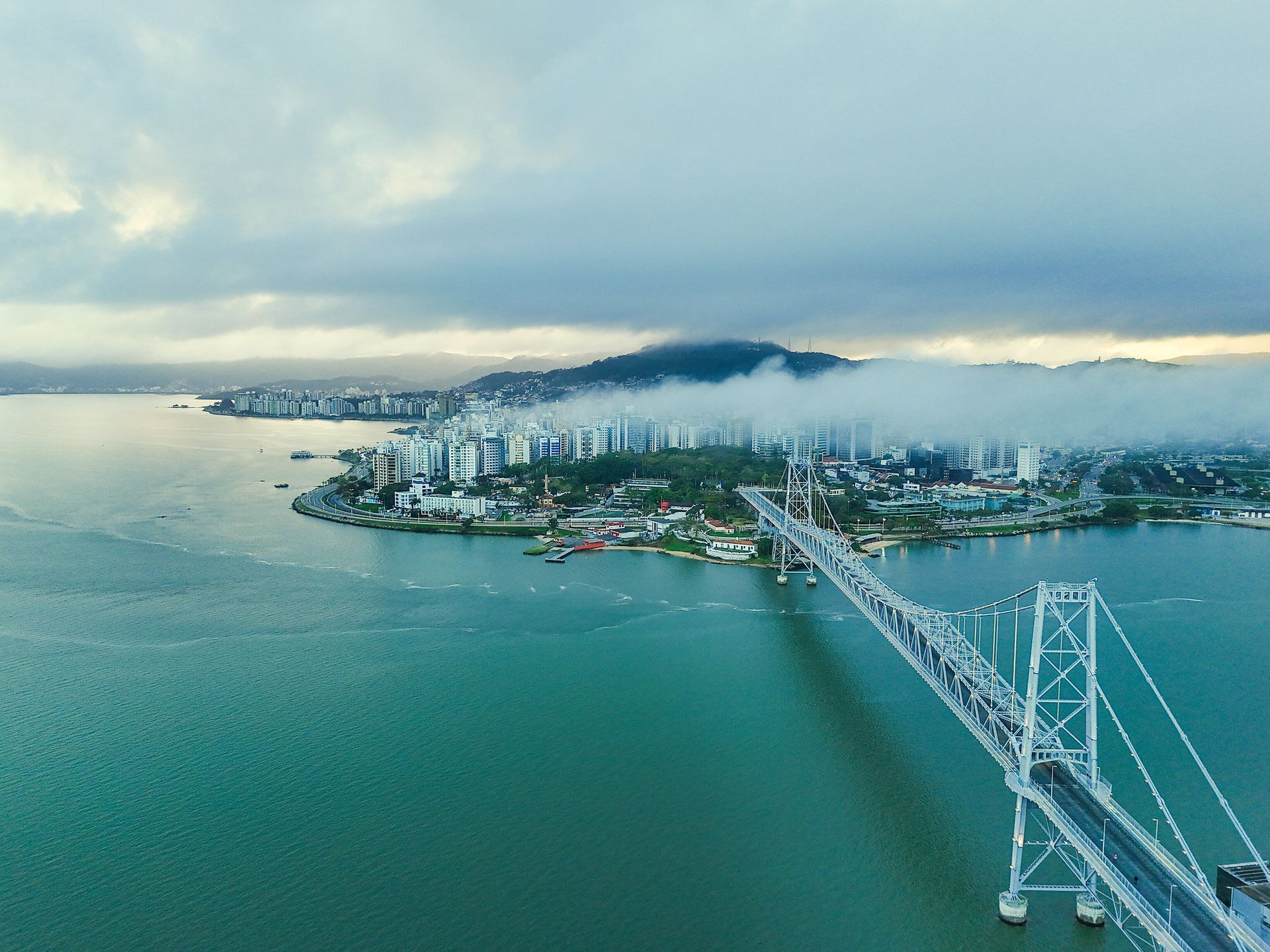 Ponte suspensa sobre águas azul-turquesa, ligando a uma cidade parcialmente encoberta por nuvens.