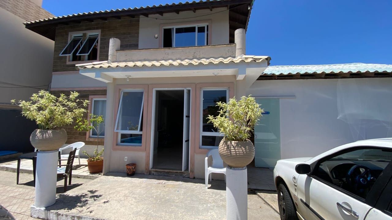 Casa de dois andares com fachada branca, plantas em vasos, céu azul e um carro estacionado na frente.