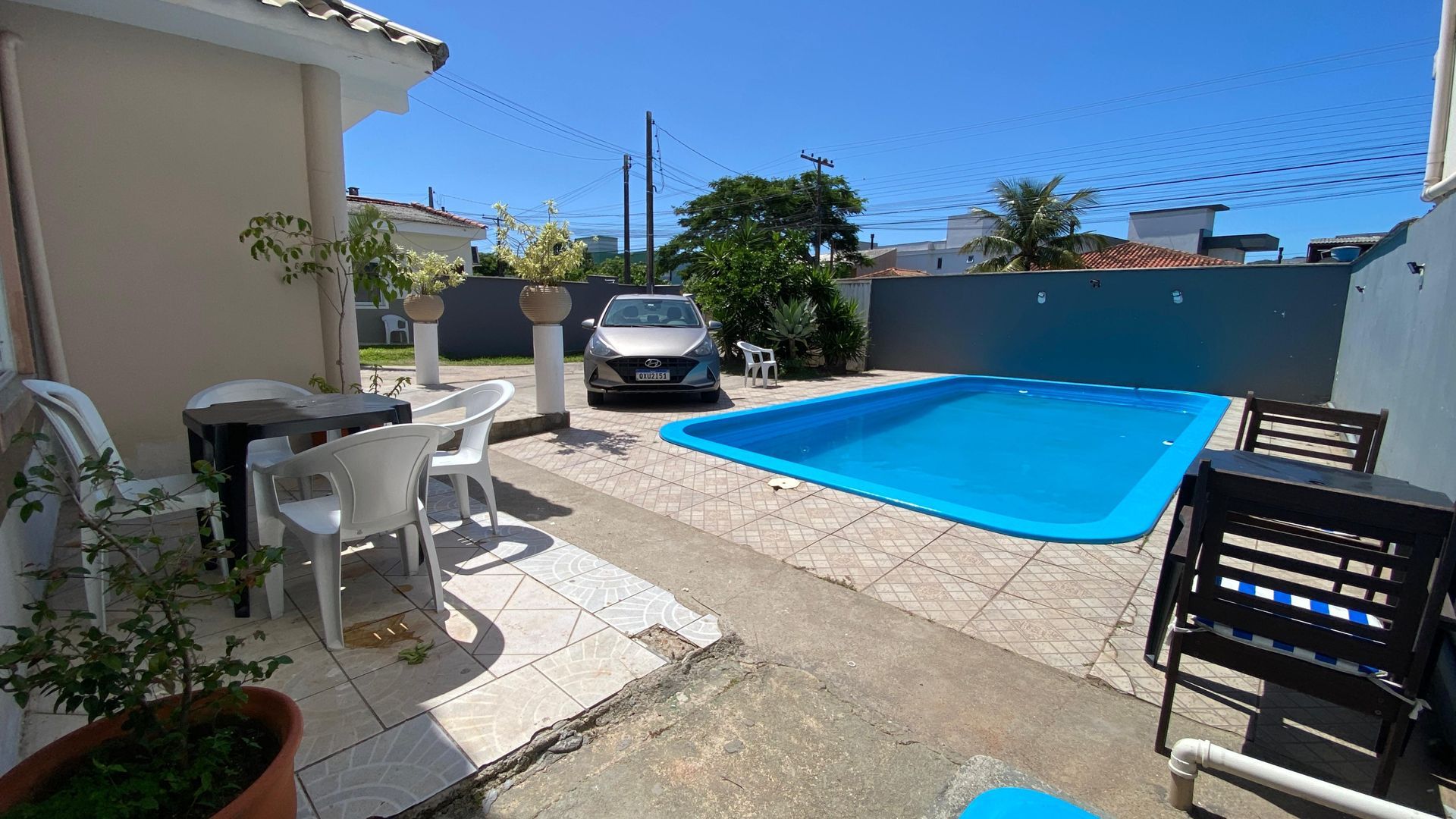 Quintal com piscina, móveis de jardim, um carro e uma casa sob um céu azul.